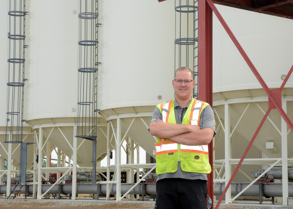 Person in a high visibility safety vest standing in front of large white storage bins at an agricultural facility.